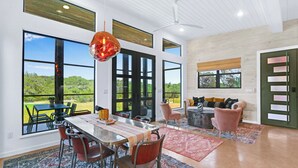 A spacious dining area with a long table and chairs, large windows offering a view of greenery, and a ceiling fan. - Wild Candy Farm - Breezeway Haus (Wimberley)