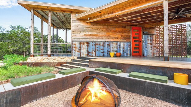 Une terrasse extérieure moderne, dotée d’un foyer, d’une plateforme en bois et d’un escalier menant à un espace couvert avec une porte rouge.