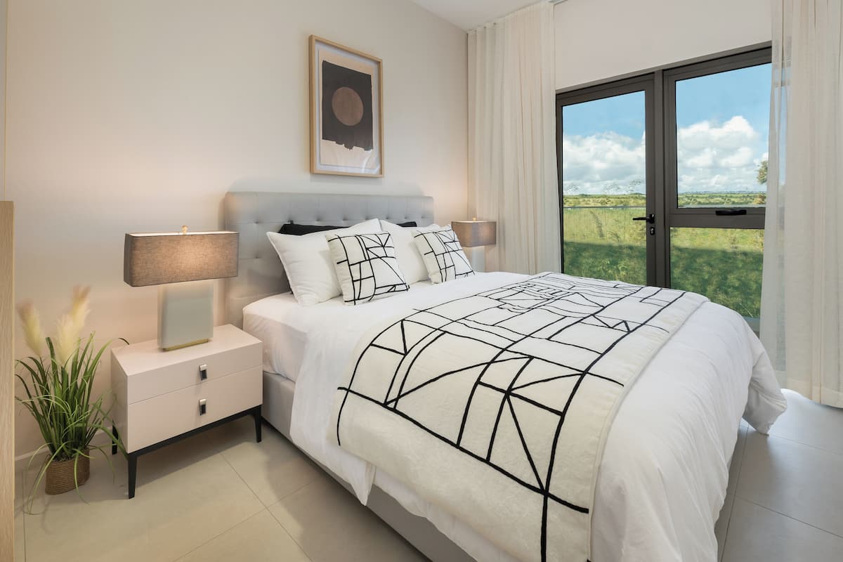 Een slaapkamer met een groot bed, twee nachtkastjes met lampen, een kunstwerk in een lijst boven het bed, en een uitzicht op groen door een schuifdeur.