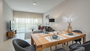 Ein modernes Wohnzimmer mit Essbereich, ausgestattet mit einem Holztisch, der mit Tellern, Gläsern und Besteck gedeckt ist. Im Wohnbereich befinden sich eine Couch, ein Fernseher und eine Stehlampe.