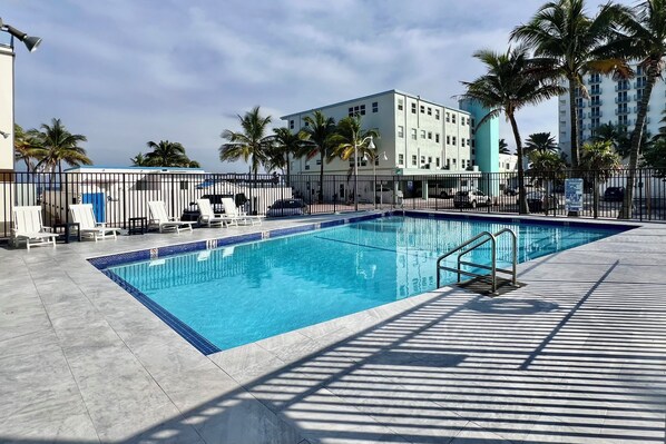 A swimming pool with lounge chairs and a fence, surrounded by palm trees and a building with multiple floors. - BRAND NEW FOR 4-1/1 BEACH VIEW-HEATED POOL-PARKING (Hollywood)