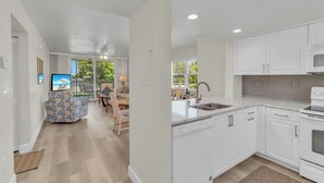 A modern kitchen with white cabinetry, a dishwasher, and a microwave. There is a dining area with a table and chairs, and a view of the outdoors through large windows.