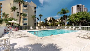 A pool area with lounge chairs and umbrellas, surrounded by palm trees and modern buildings.