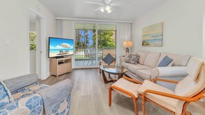 A living room with a television, a sofa, a coffee table, and a chair. There is a balcony with a view of palm trees and a beach.