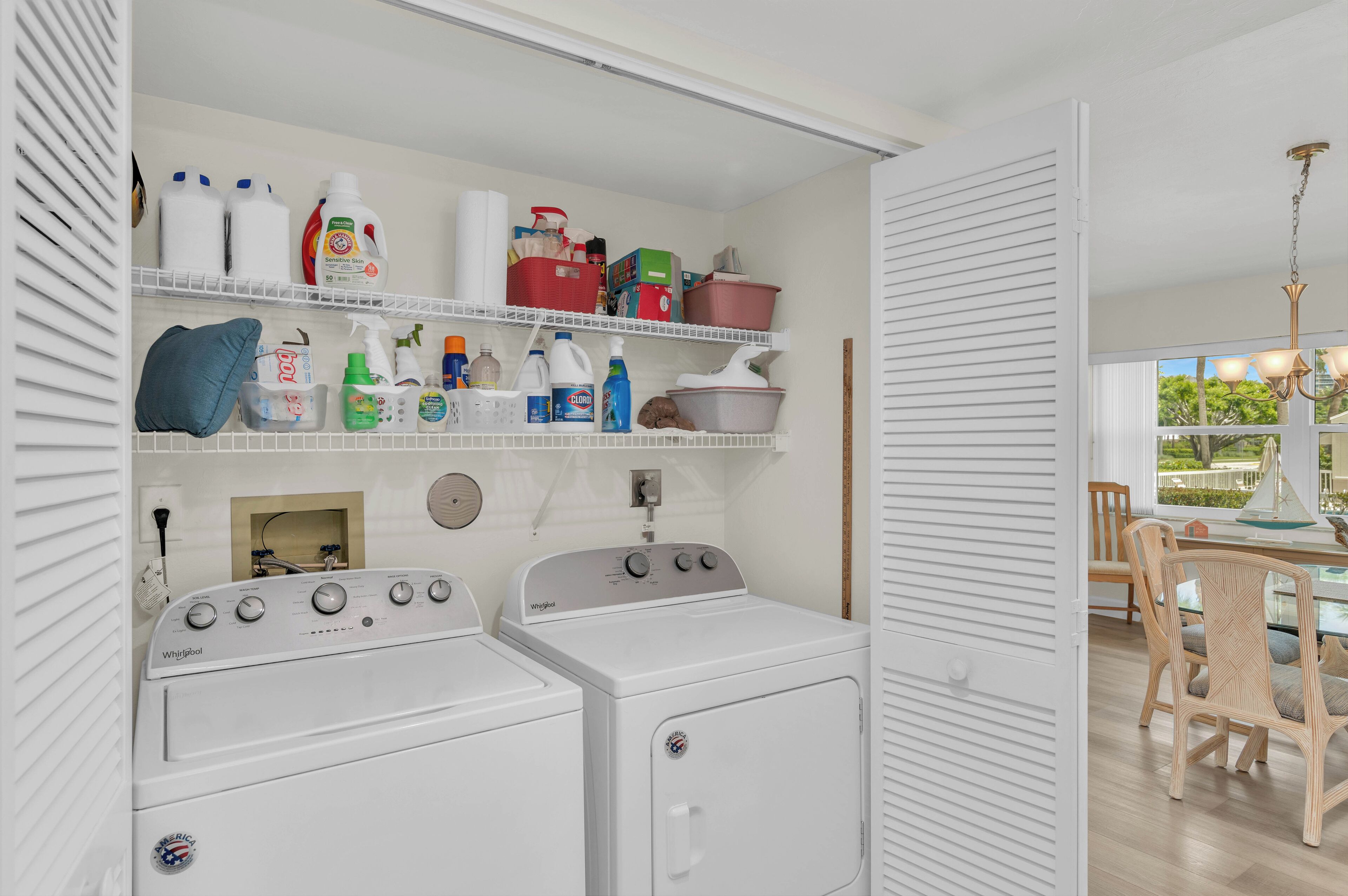 A laundry area with two white Whirlpool washers, shelves with cleaning supplies, and a dining area with wicker chairs and a table.