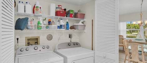 A laundry area with two white Whirlpool washers, shelves with cleaning supplies, and a dining area with wicker chairs and a table.