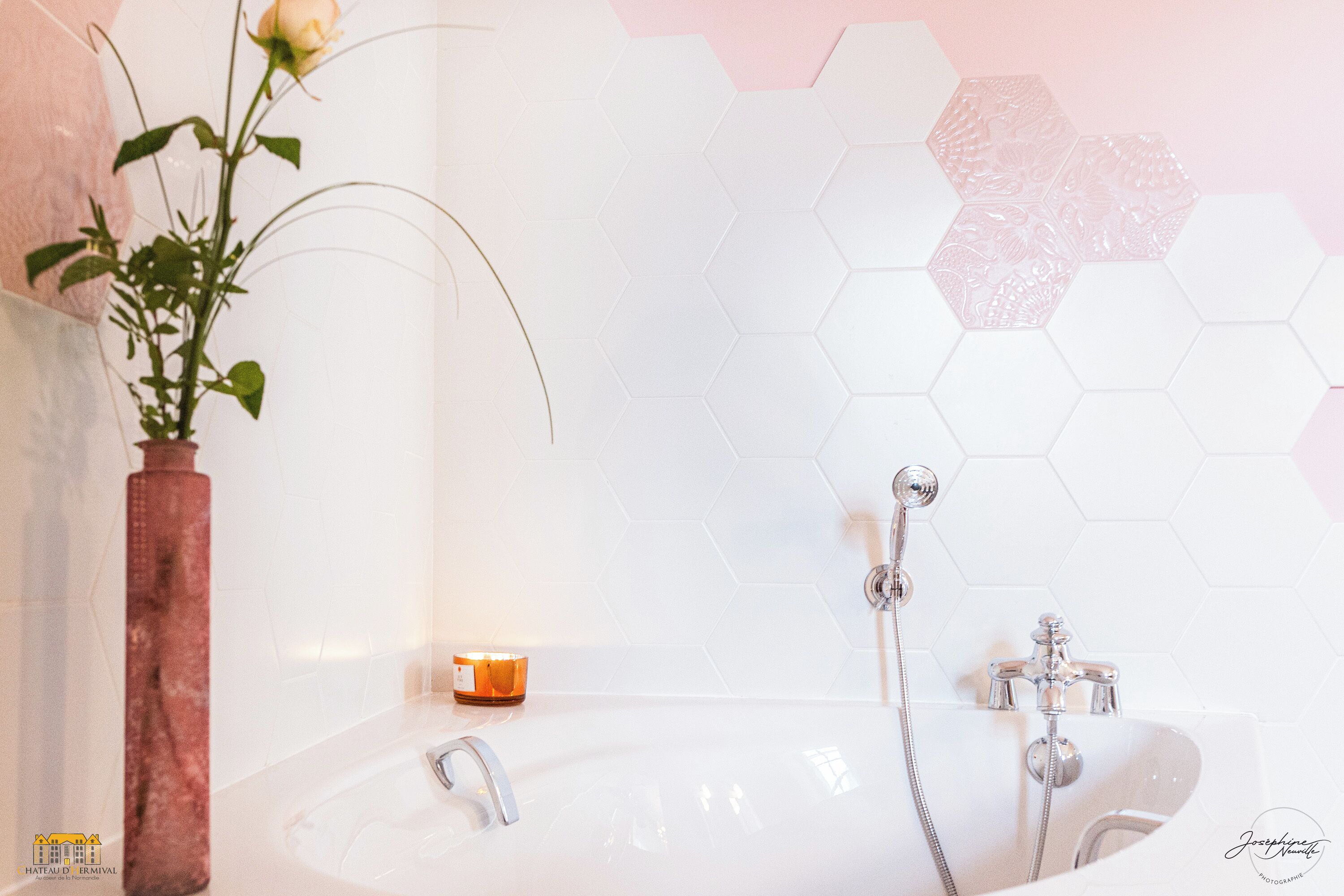 Une salle de bain avec une baignoire blanche, une rose dans un vase et une bougie posée sur le bord.
