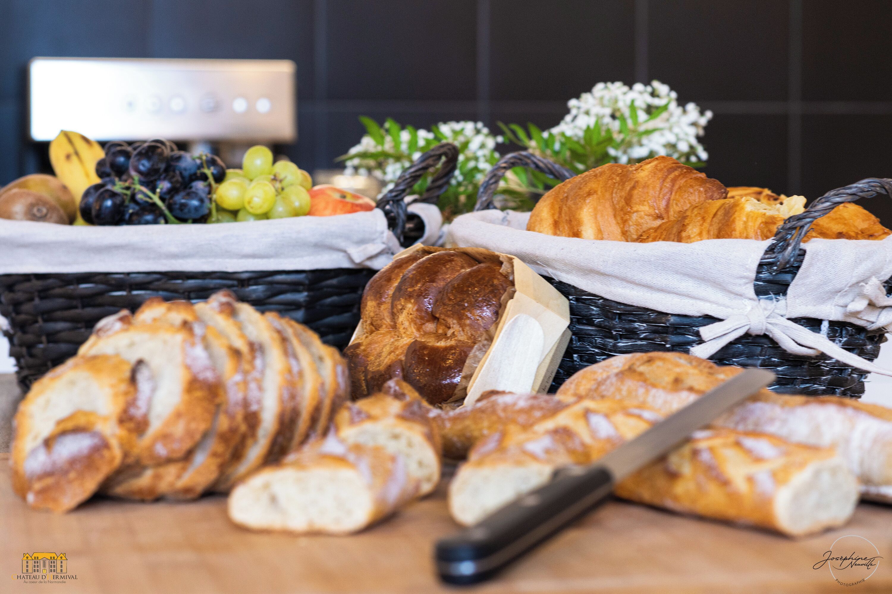 A variety of bread and fruits on a wooden table with a knife, in front of a basket tied with a ribbon.