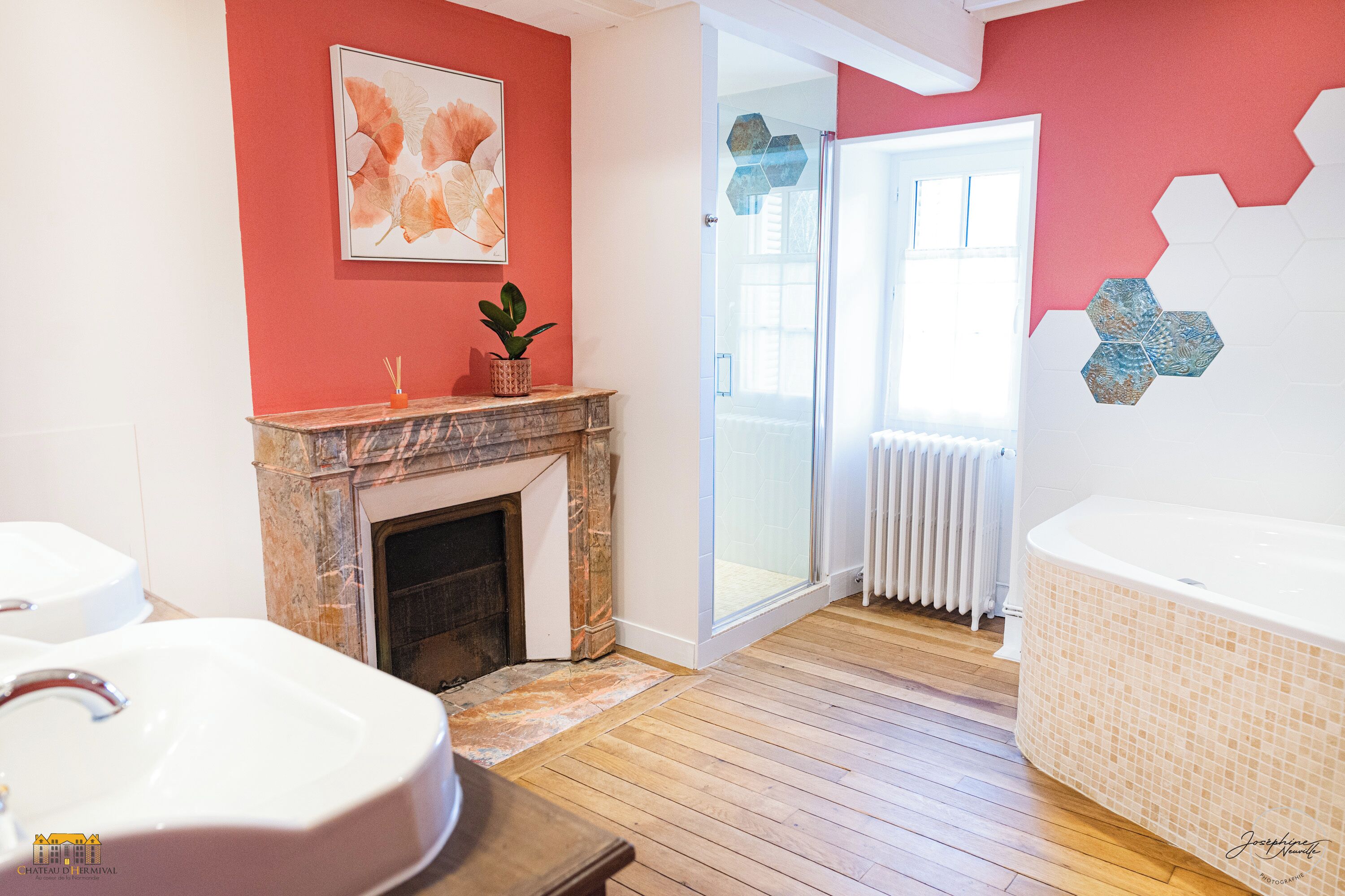 A bathroom with a wooden floor, a white bathtub, a marble fireplace, and a red accent wall.