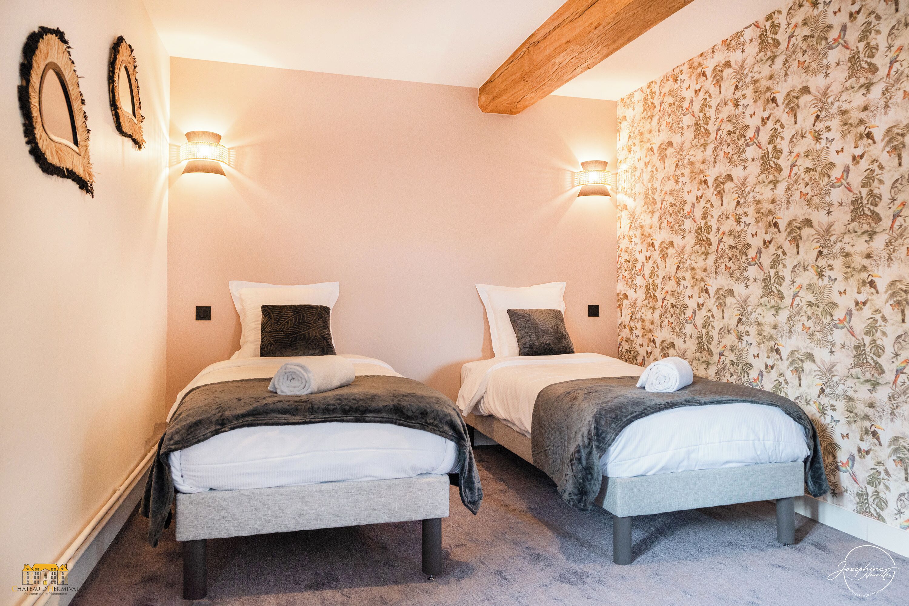 A hotel room with two beds, each with a pillow and a towel, a wooden ceiling beam, and a floral wallpaper on one wall.