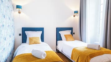 A hotel room with two beds, each with a blue headboard, white bedding, and a mustard-colored throw blanket. There are wall-mounted lamps on either side of the beds, and a window with curtains on the right side.