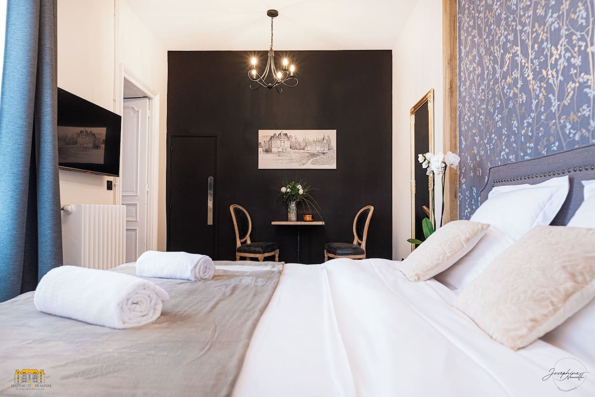 A hotel room with a bed, two chairs, a small table with a vase, and a chandelier.