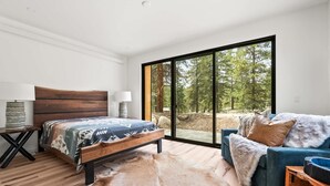 Un dormitorio con cama, sofá, ventana grande con vistas al bosque y suelo de madera.