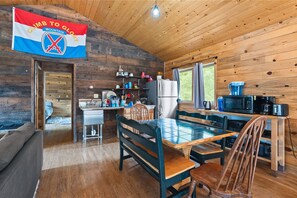 A cabin interior with a wooden interior, a dining area with a table and chairs, a kitchen with appliances, and a flag on the wall. - Hwy 91 Log Cabin — 2BR/2BA, Fireplace, Mtn Views, Sleeps 4 (Leadville)