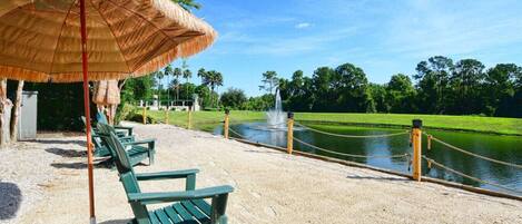 A beach area with a thatched umbrella, green lounge chairs, a sandy walkway, and a water body with a fountain.