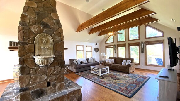 A spacious living room with a stone fireplace, wooden beams, large windows, and a cozy seating area.