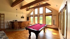A spacious room with a pool table, wooden flooring, and large windows offering a view of the outdoors.