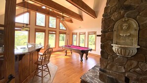 A spacious room with a pool table, wooden ceiling beams, large windows, and a stone fireplace.