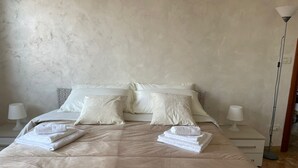 A neatly made bed with a beige duvet, two pillows, and folded towels. There are two bedside tables with lamps and a floor lamp. - Like At Home Venice Mestre, bright, family and private parking place. (Venezia)