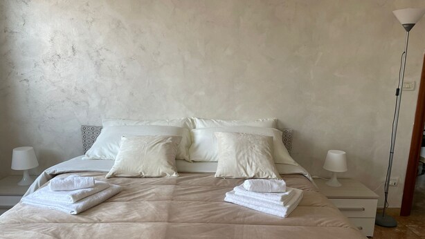 A neatly made bed with a beige duvet, two pillows, and folded towels. There are two bedside tables with lamps and a floor lamp. - Like At Home Venice Mestre, bright, family and private parking place. (Venezia)