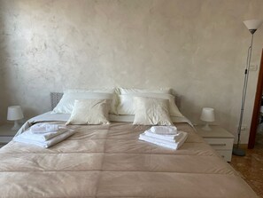 A neatly made bed with a beige duvet, two pillows, and folded towels. There are two bedside tables with lamps and a floor lamp.