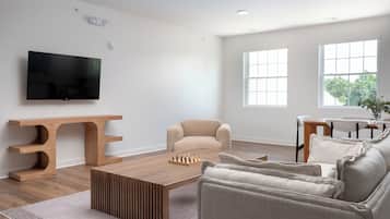 A modern living room with a flat-screen TV, a wooden entertainment unit, a beige sofa, a coffee table, and large windows.