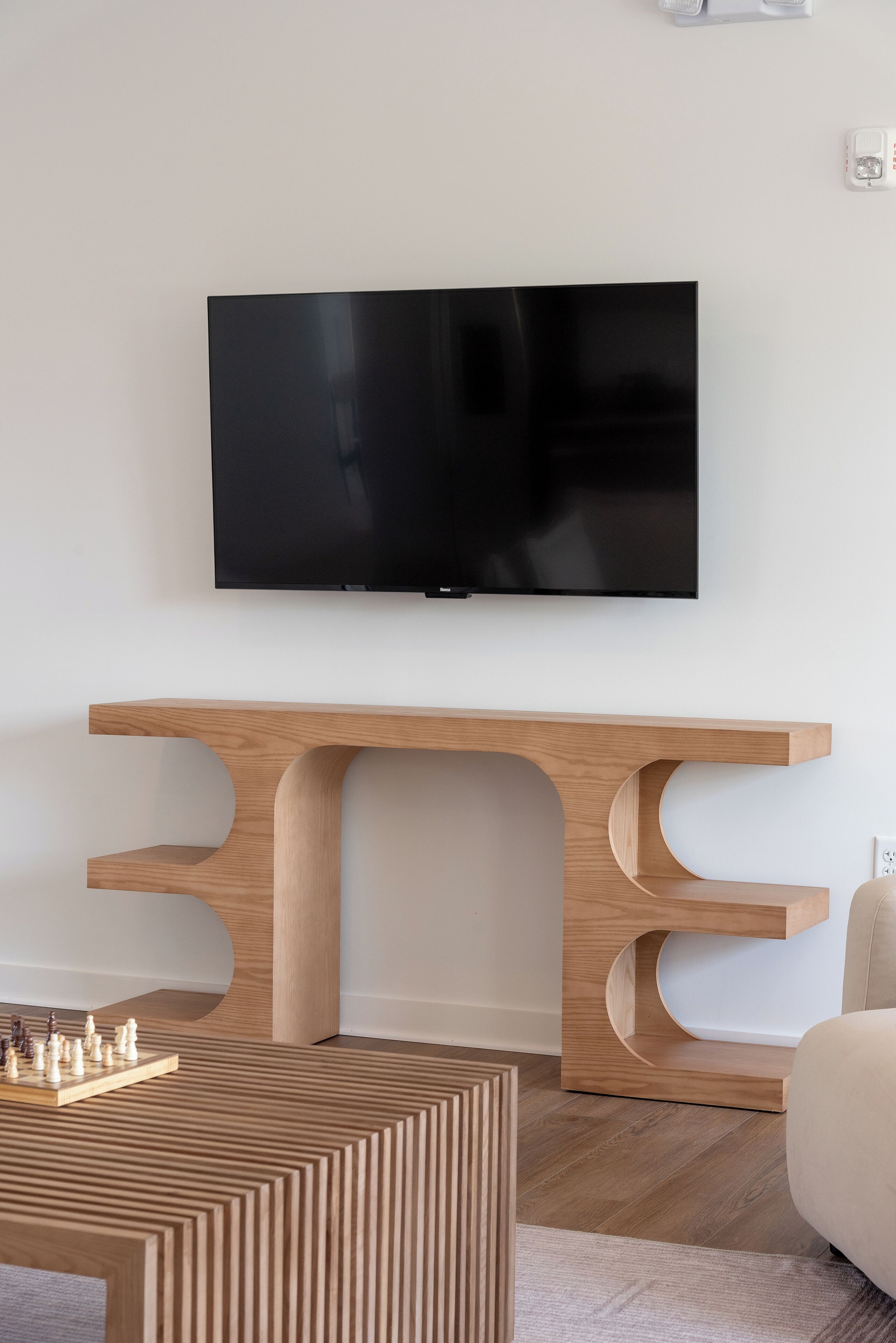 A modern living room with a flat-screen TV mounted on the wall, a wooden console table with a chessboard, and a patterned ottoman.