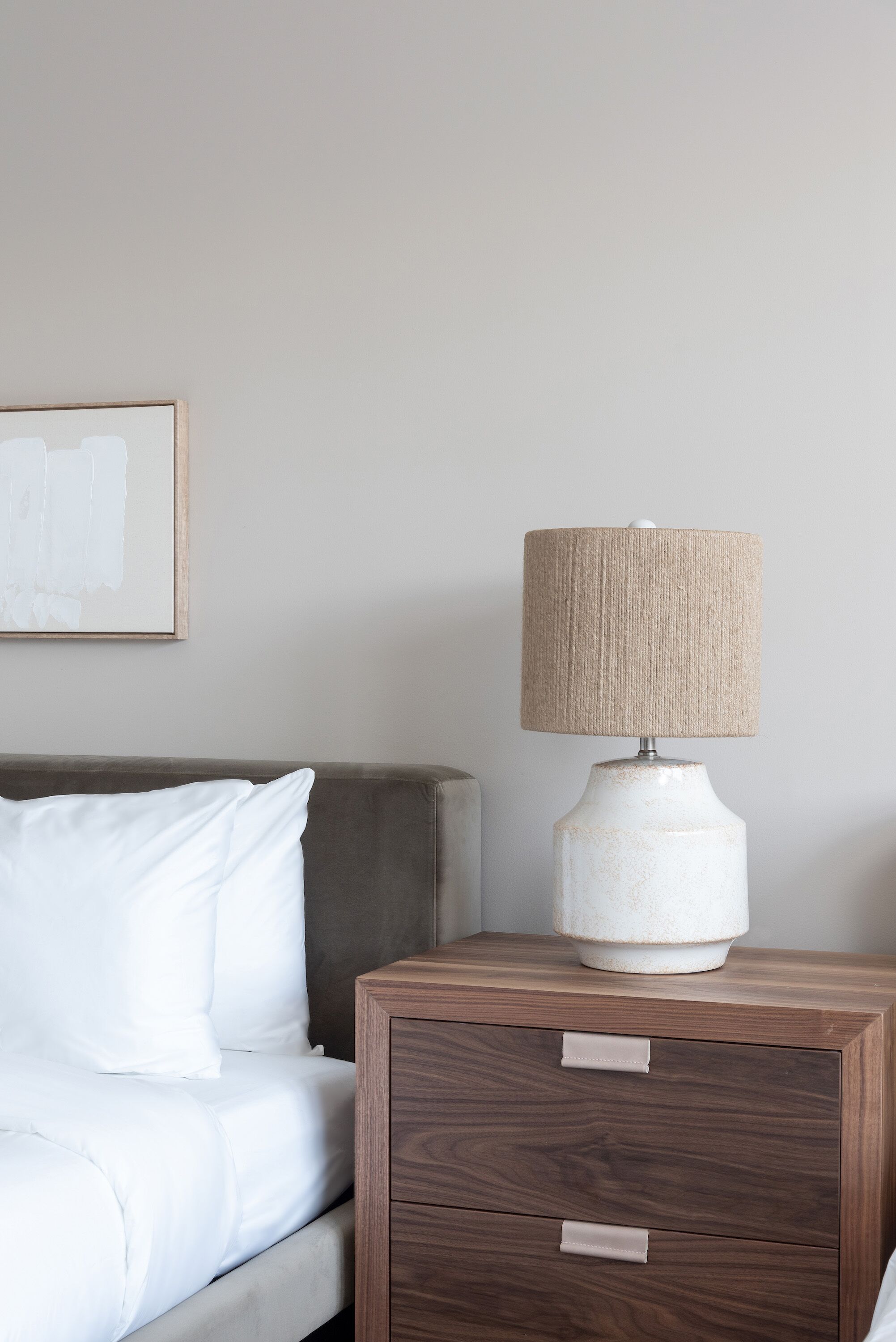 Ein ordentlich bezogenes Bett mit weißer Bettwäsche, ein Nachttisch aus Holz mit Lampe und ein gerahmtes Bild an der Wand.