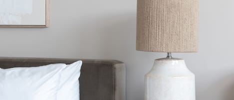 Ein ordentlich bezogenes Bett mit weißer Bettwäsche, ein Nachttisch aus Holz mit Lampe und ein gerahmtes Bild an der Wand.