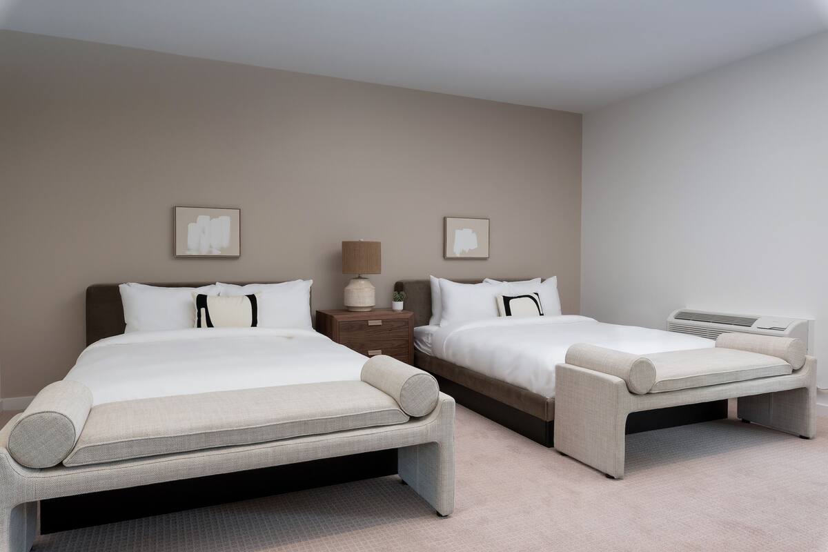 Habitación de hotel con dos camas, una mesita de noche, una lámpara y dos cuadros en la pared.