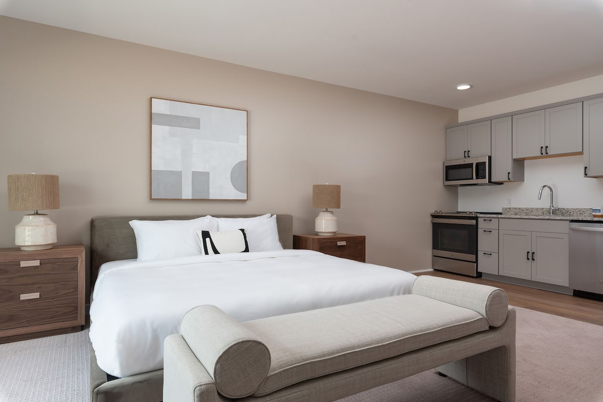 Una habitación de hotel moderna con una cama grande, lámparas de noche, un escritorio pequeño y una kitchenette.