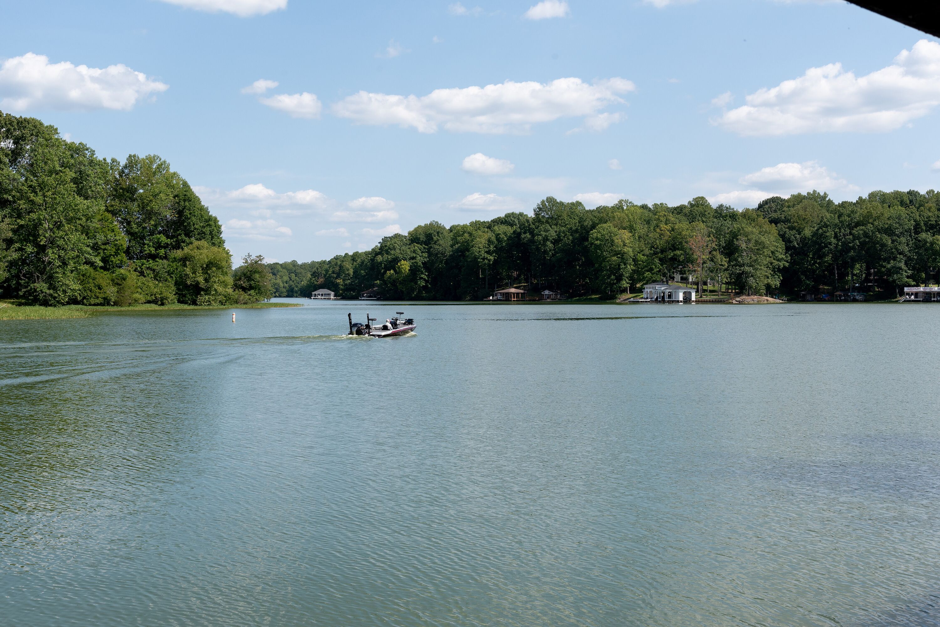 A calm lake with a boat, surrounded by dense forest and several buildings on the shore.