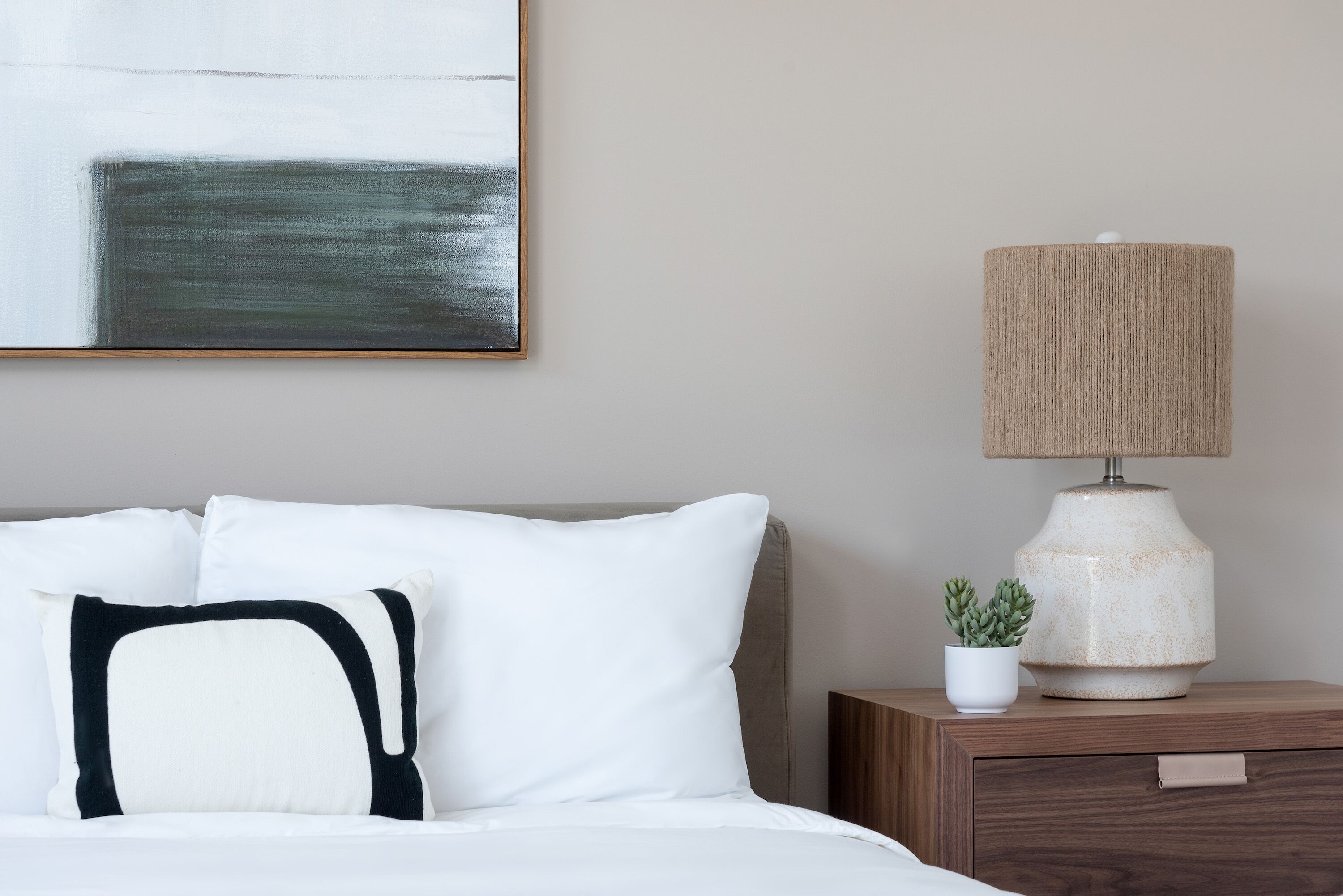 Una cama bien hecha con almohadas blancas y negras, una pared color beige, un cuadro abstracto enmarcado, una mesita de noche de madera con una lámpara y una pequeña planta en maceta.