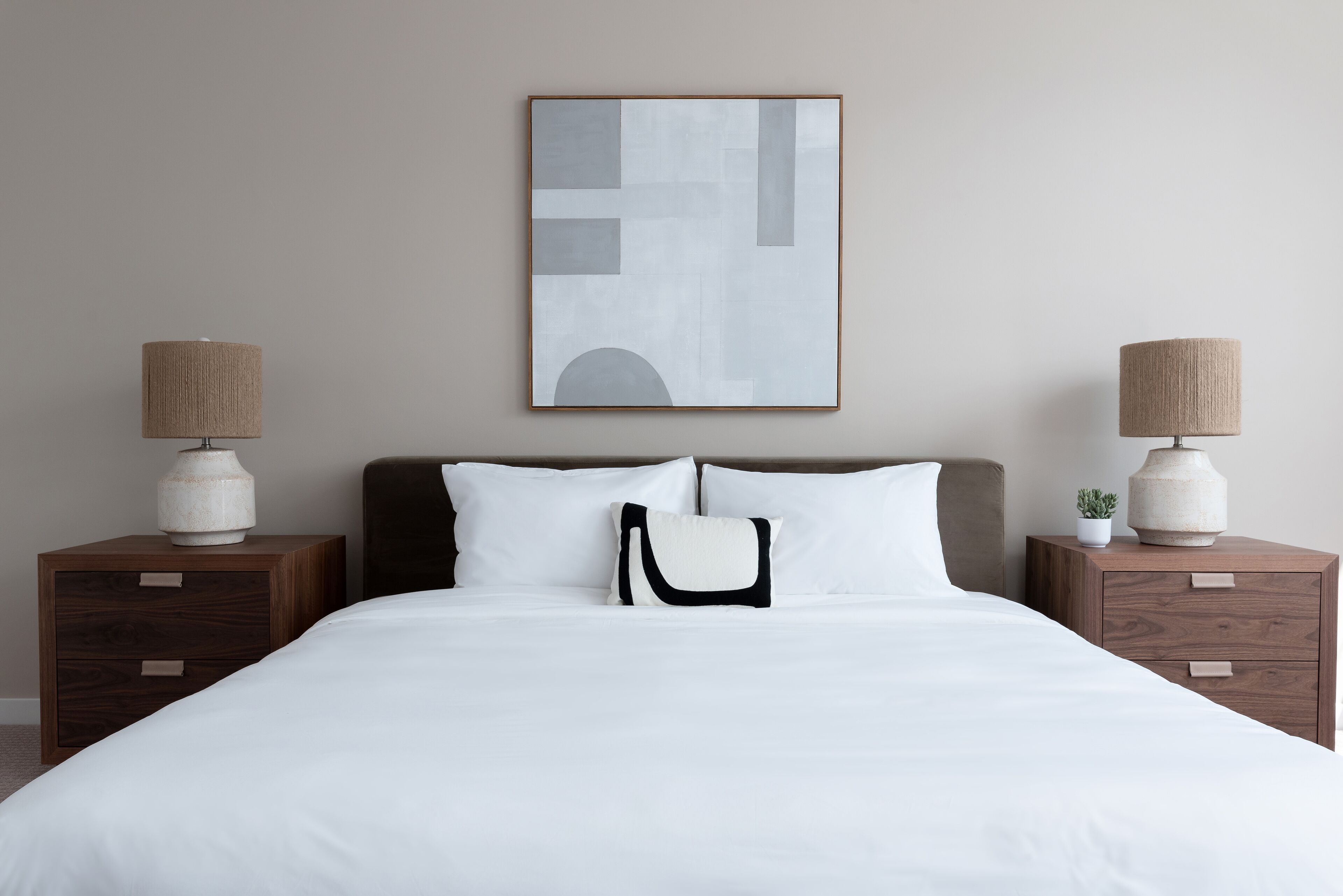 Een keurig opgemaakt bed met witte lakens, twee nachtkastjes met lampen, en een lijst met abstracte kunst aan de muur.