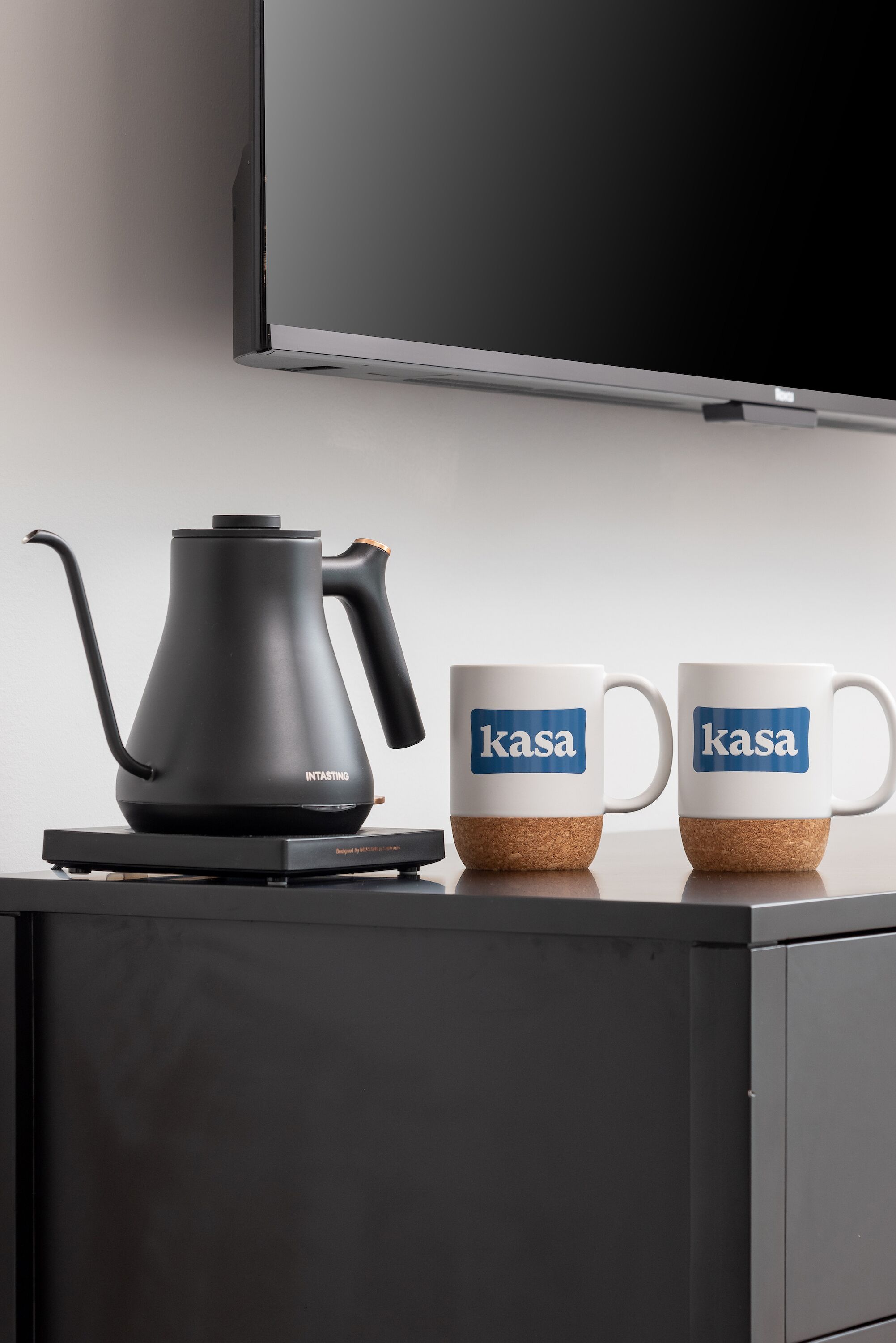 En sort elkedel og to hvide kopper med "kasa" på, placeret på en sort overflade med et fjernsyn over.