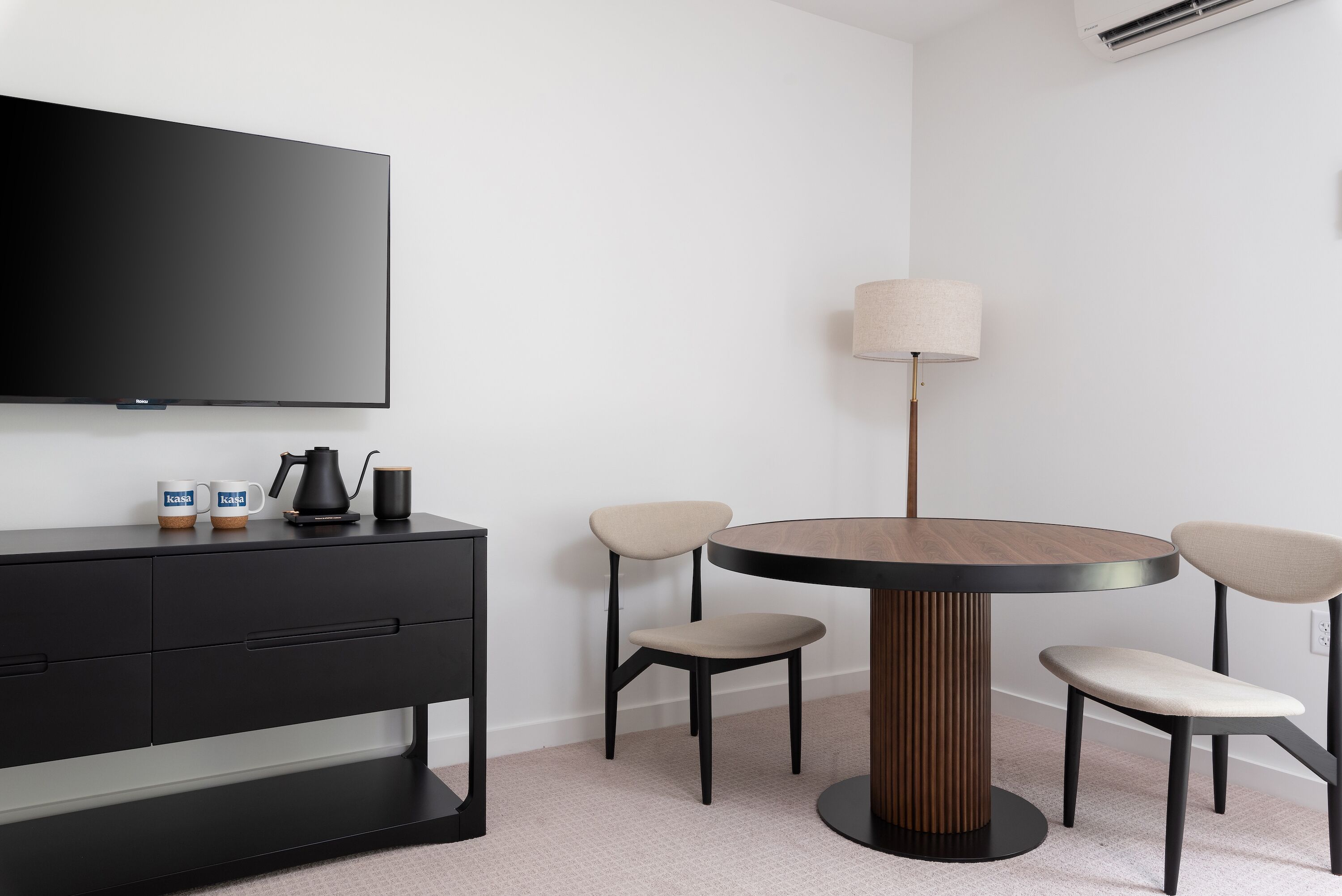 Una habitación moderna con un televisor de pantalla plana montado en la pared, una consola negra con dos tazas, una cafetera y una lámpara. Cuatro sillas rodean una mesa redonda de madera.