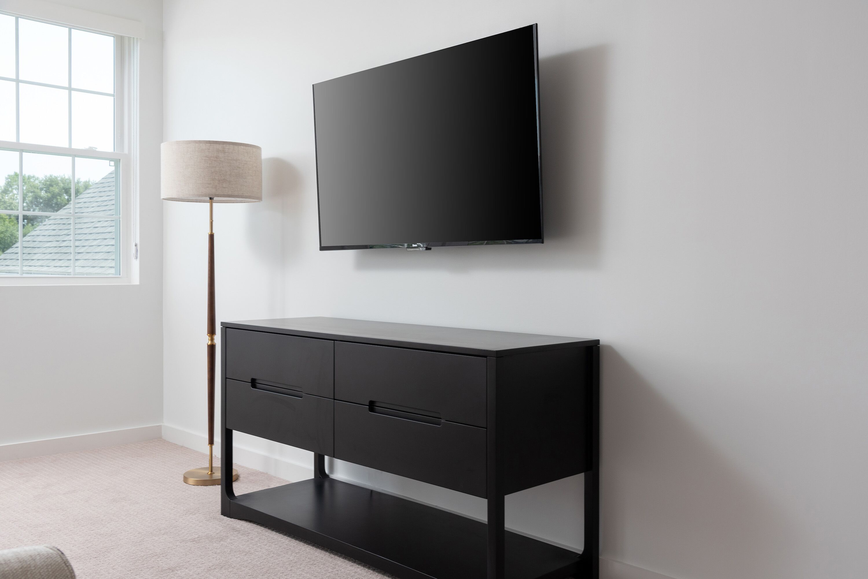A modern room with a black TV on the wall, a black console table with drawers, a floor lamp, and a window with a view of trees.