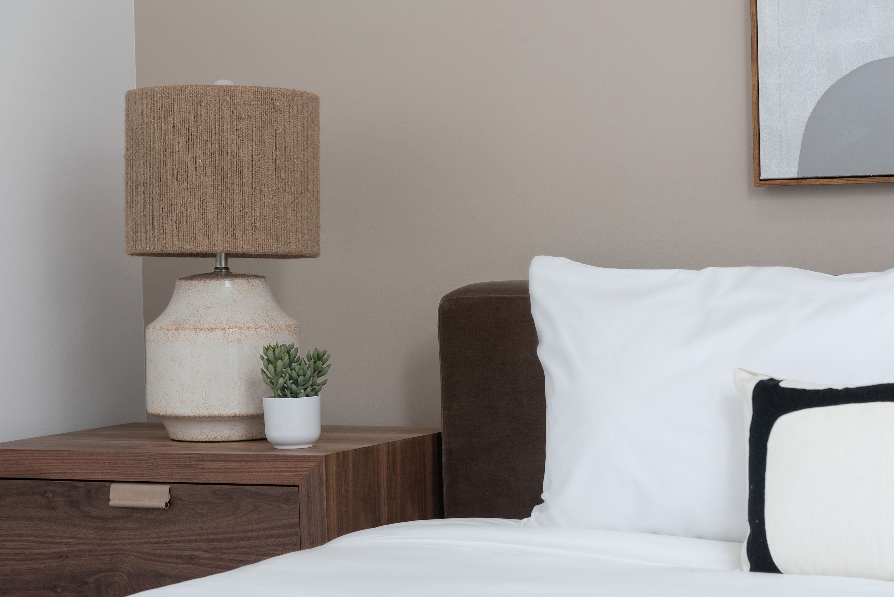 Ein ordentlich bezogenes Bett mit weißen und schwarzen Kissen, ein Nachttisch aus Holz mit Lampe und kleiner Topfpflanze sowie ein gerahmtes Bild an der Wand.