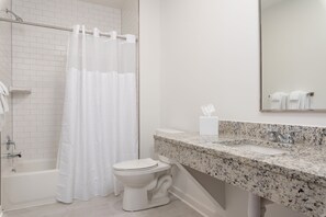 Une salle de bain avec des toilettes blanches, une douche avec un rideau blanc, un lavabo sur un plan de travail en granit et un miroir au-dessus du lavabo.