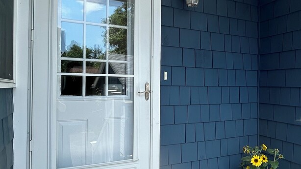 Eine weiße Tür mit Fensterglas, eine dunkelblaue Außenwand, ein Blumentopf mit gelben Blüten und eine Fußmatte mit der Aufschrift 'HOME'.