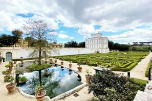 A formal garden with a central pond, manicured hedges, and a classical building in the background.