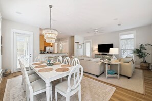 Un coin repas avec une table et des chaises blanches, un lustre et une cuisine en arrière-plan.