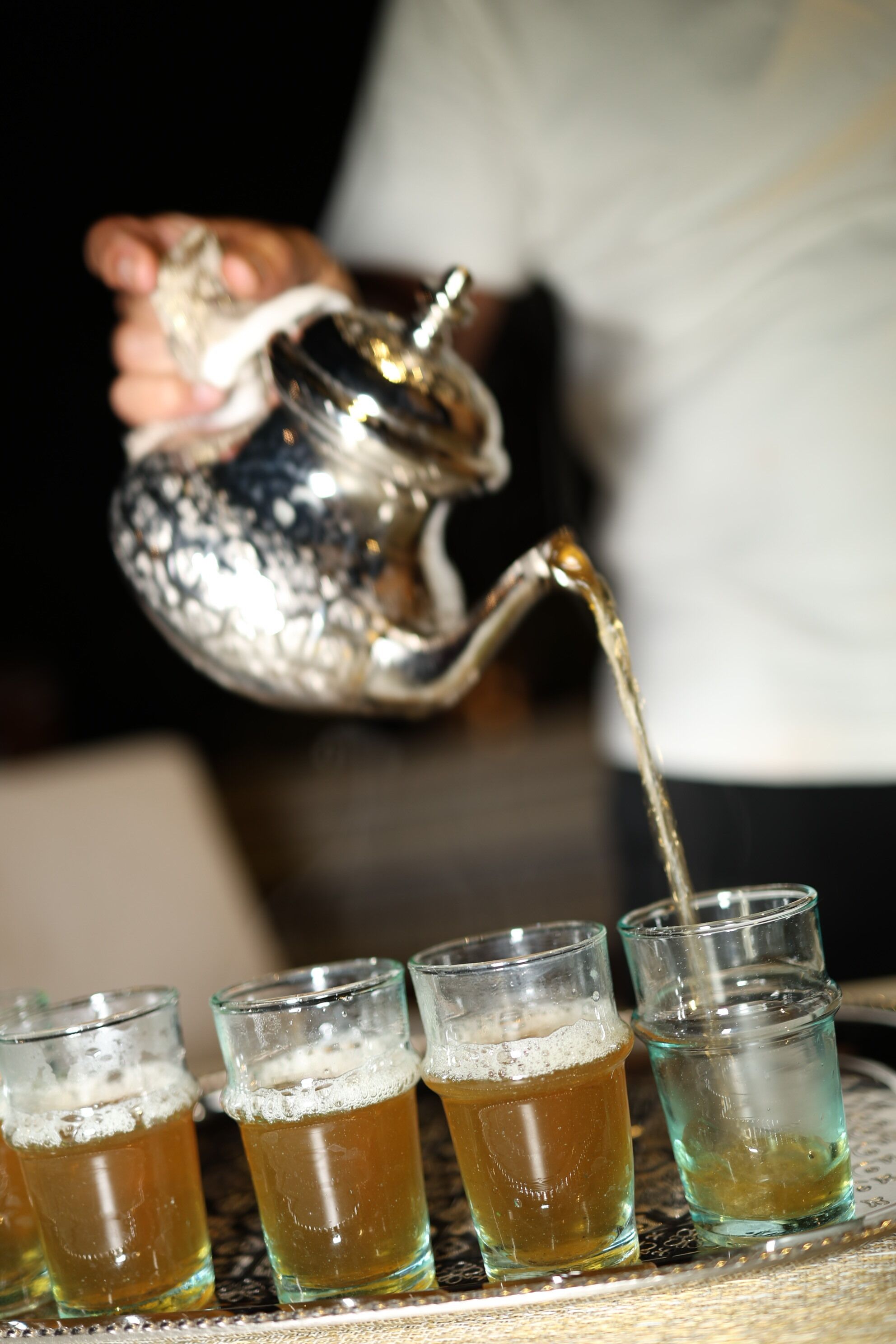 A hand pouring liquid from a silver teapot into four glasses.
