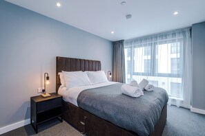 A neatly made bed with white linens and a gray blanket, a bedside table with a lamp, and a window with sheer curtains. - Hilltop Serviced Apartments-Sheffield (Sheffield)
