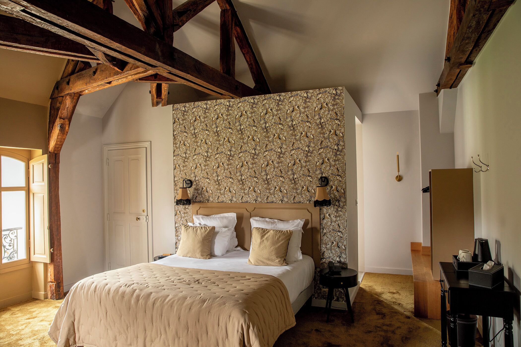 Quarto com cama grande, vigas de madeira, parede estampada e janela com cortinas.