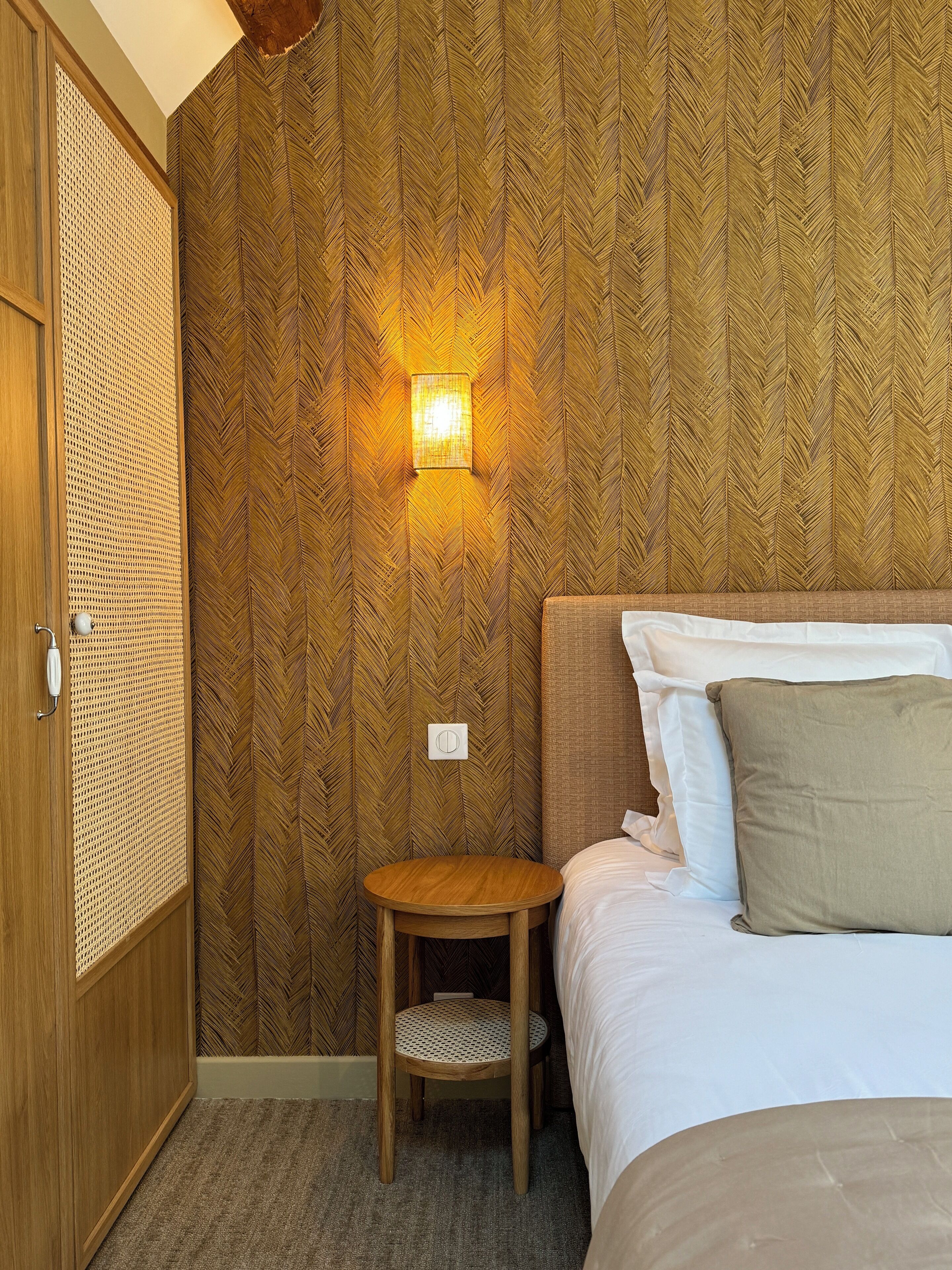 Une chambre d’hôtel avec un papier peint à motifs chevrons, une table ronde en bois, un lit avec une literie blanche et beige, et un luminaire fixé au mur.