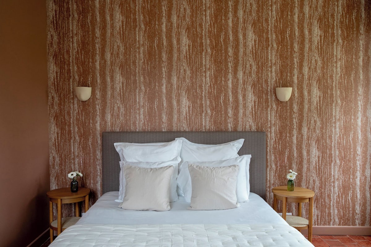 Een net opgemaakt bed met witte lakens en kussens, twee nachtkastjes met kleine bloemenvaasjes, en een gestructureerde wand achter het bed.