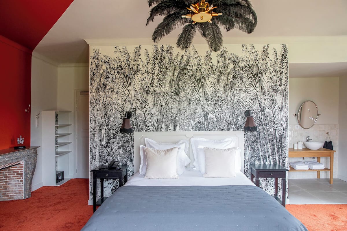 Een slaapkamer met een bed, een nachtkastje, een houten bijzettafeltje, een rood tapijt, een wit hoofdeinde, een muur met een zwart-wit patroon, een plafondventilator en een badkamer met een wastafel en spiegel.