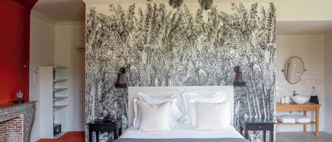 Un dormitorio con cama, mesita de noche, una mesita auxiliar de madera, alfombra roja, cabecera blanca, pared con diseño blanco y negro, ventilador de techo, y un baño con lavamanos y espejo.