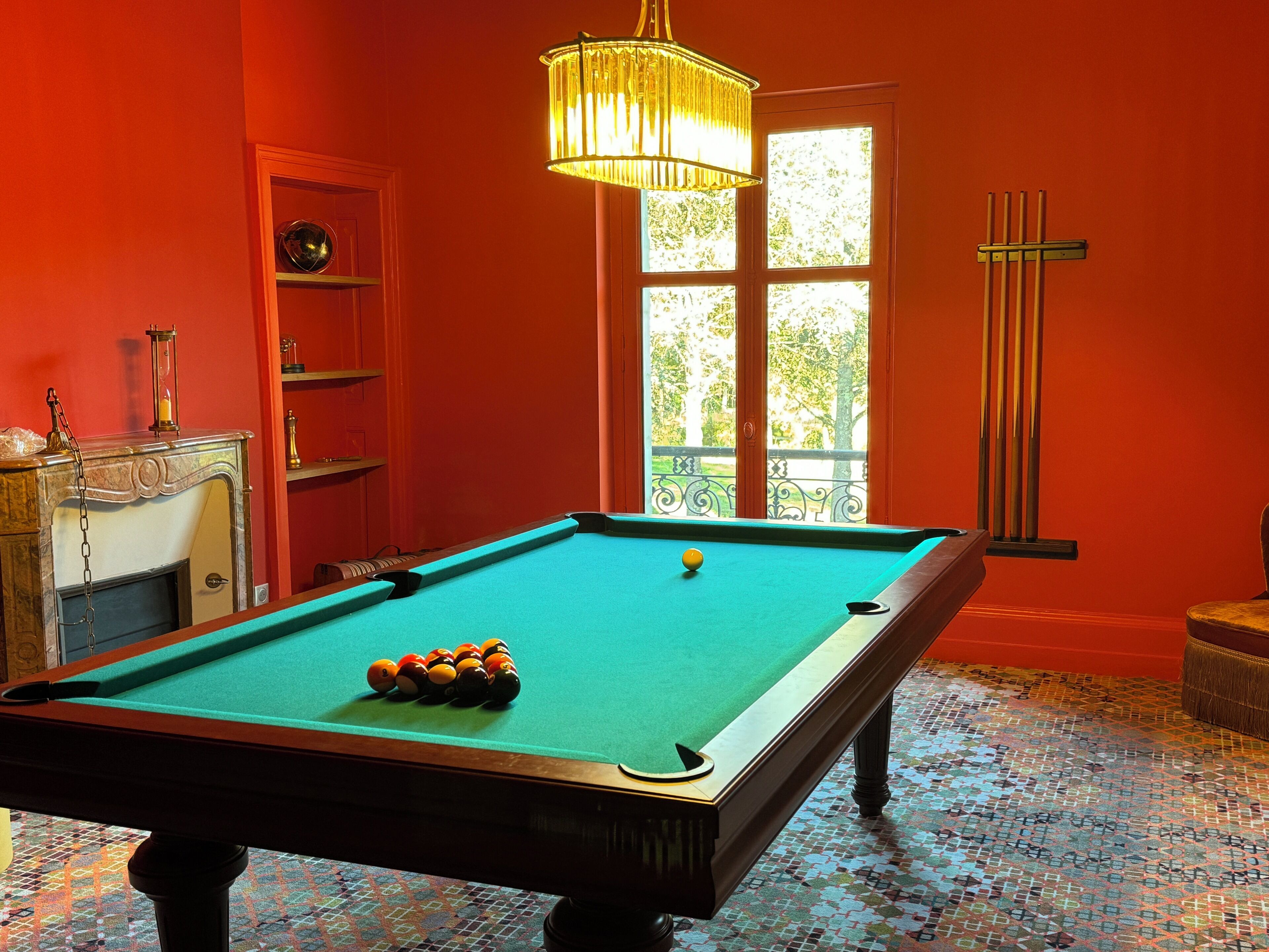 A pool table in a room with red walls, a chandelier, and a window with a view of greenery.